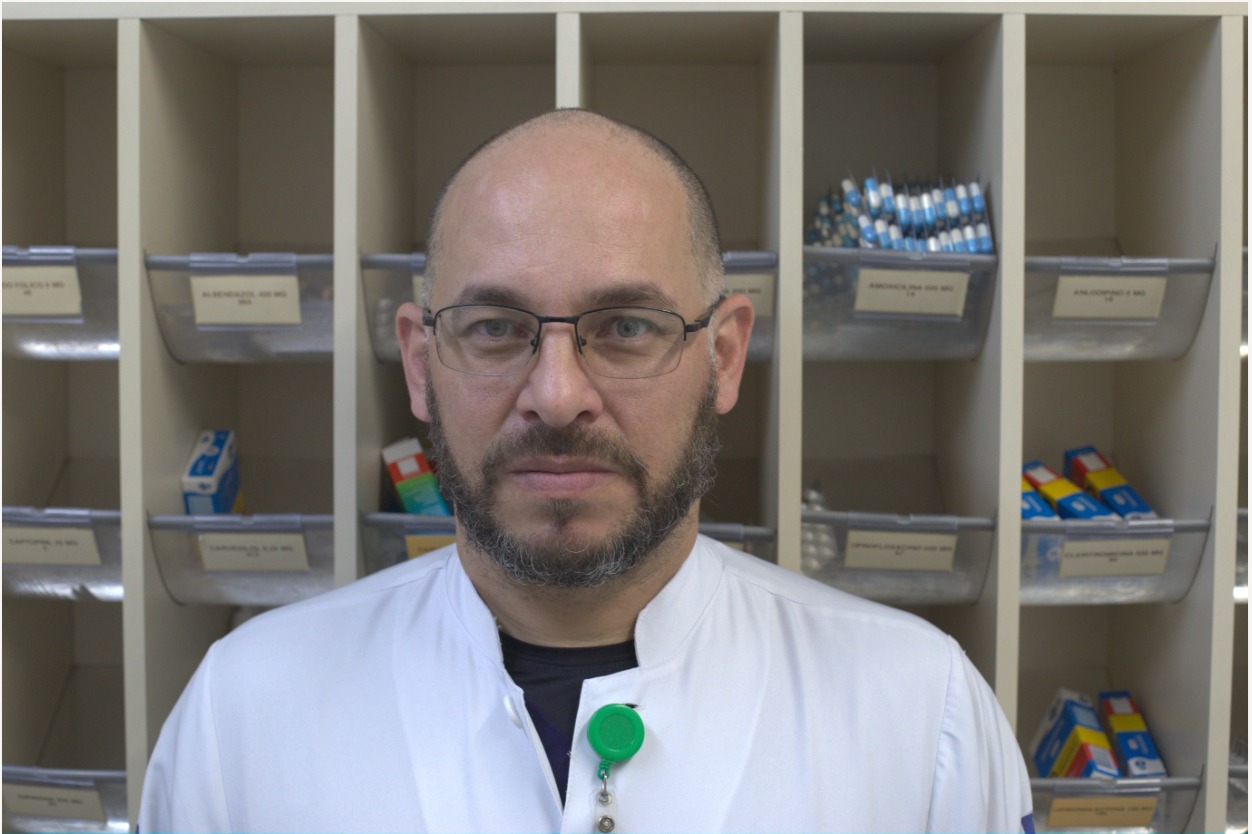 Na imagem, aparece Antônio e, ao fundo, uma parede com medicamentos. Ele é um homem pardo, de 48 anos, careca, com barba e usa óculos de grau, e jaleco branco.